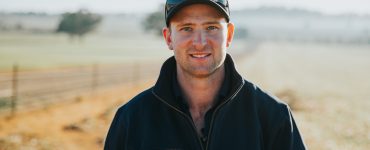 Man smiling in misty paddock, wearing dark jumper and cap.